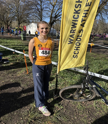 Photograph of a young athlete standing beside a yellow vertical banner reading "Warwickshire Schools Athletics" at an outdoor event with grass and trees in the background. The athlete wears a yellow vest with a race number 484 and dark trousers, with a muddy bicycle resting against the banner stand