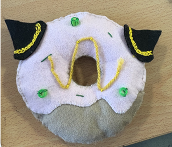 Photograph of a handmade plush donut with light pink frosting and beige base, decorated with black felt ears featuring yellow embroidered details. The donut also has green sequin accents and yellow embroidered squiggles on frosting, placed on a wooden surface 