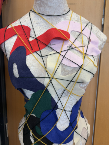 A photograph of a dressmaker's mannequin decorated with multicoloured felt shapes and crisscrossing yellow and black yarn strands. The abstract arrangement creates a layered, textured design with red, pink, navy, green, blue, and cream felt pieces contrasting against the neutral mannequin.