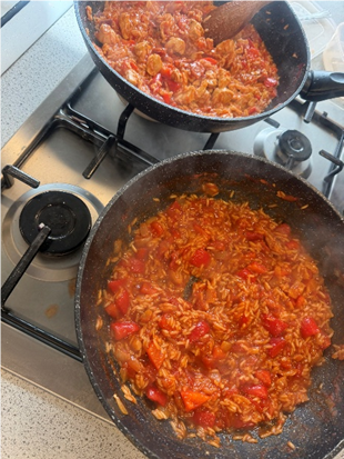 Photograph showing two frying pans on a gas stove, each containing a tomato-based rice dish with visible chunks of red peppers and pieces of meat or seafood. Steam rising from pans indicates cooking in progress, with a wooden spatula resting in one pan and a speckled countertop surrounding the stove