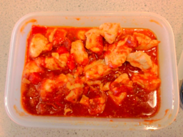 a rectangular plastic container filled with cooked chicken pieces in a red tomato-based sauce. Chicken chunks are coated evenly with sauce, showing a rich, thick texture with visible tomato bits.