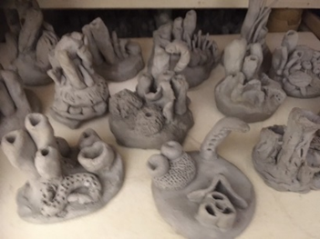 Photograph of multiple small, grey clay sculptures arranged on a flat surface, each featuring abstract, organic shapes and textured details. Sculptures vary in form, including tubular, spiral, and rounded elements, showcasing creative exploration of clay modeling technique