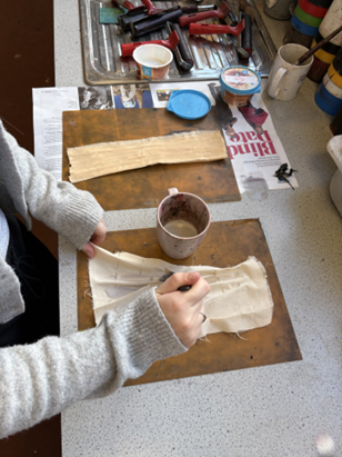 Photograph of a workspace showing a person painting or applying a substance to a piece of fabric on a brown mat, with another fabric strip and various tools and containers nearby. The scene highlights a crafting or art project involving fabric manipulation, with visible paintbrushes, cups, and a newspaper used as a protective surface.