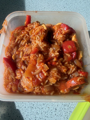 Photograph of a plastic container filled with a homemade rice dish mixed with red bell peppers and chunks of meat in a tomato-based sauce. The dish appears saucy and well-cooked, with visible pieces of vegetables and meat evenly distributed throughout the rice.