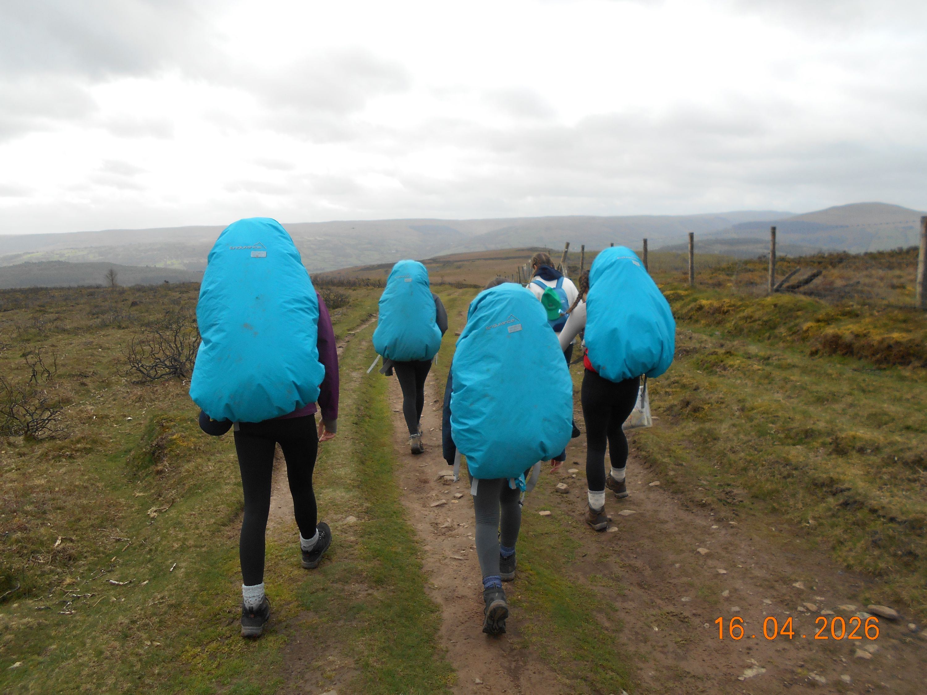 Photograph of five hikers walking along a dirt path in a rural, hilly landscape under a cloudy sky. Each hiker carries a large backpack covered with a bright blue rain cove