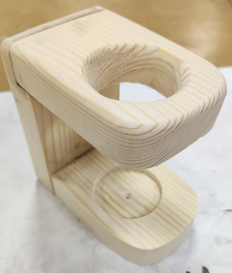 Photograph of a small wooden model resembling a simplified coffee machine with a circular cutout on top and a round indentation on the base. The model is made from light-coloured wood with visible grain patterns and smooth, rounded edges,