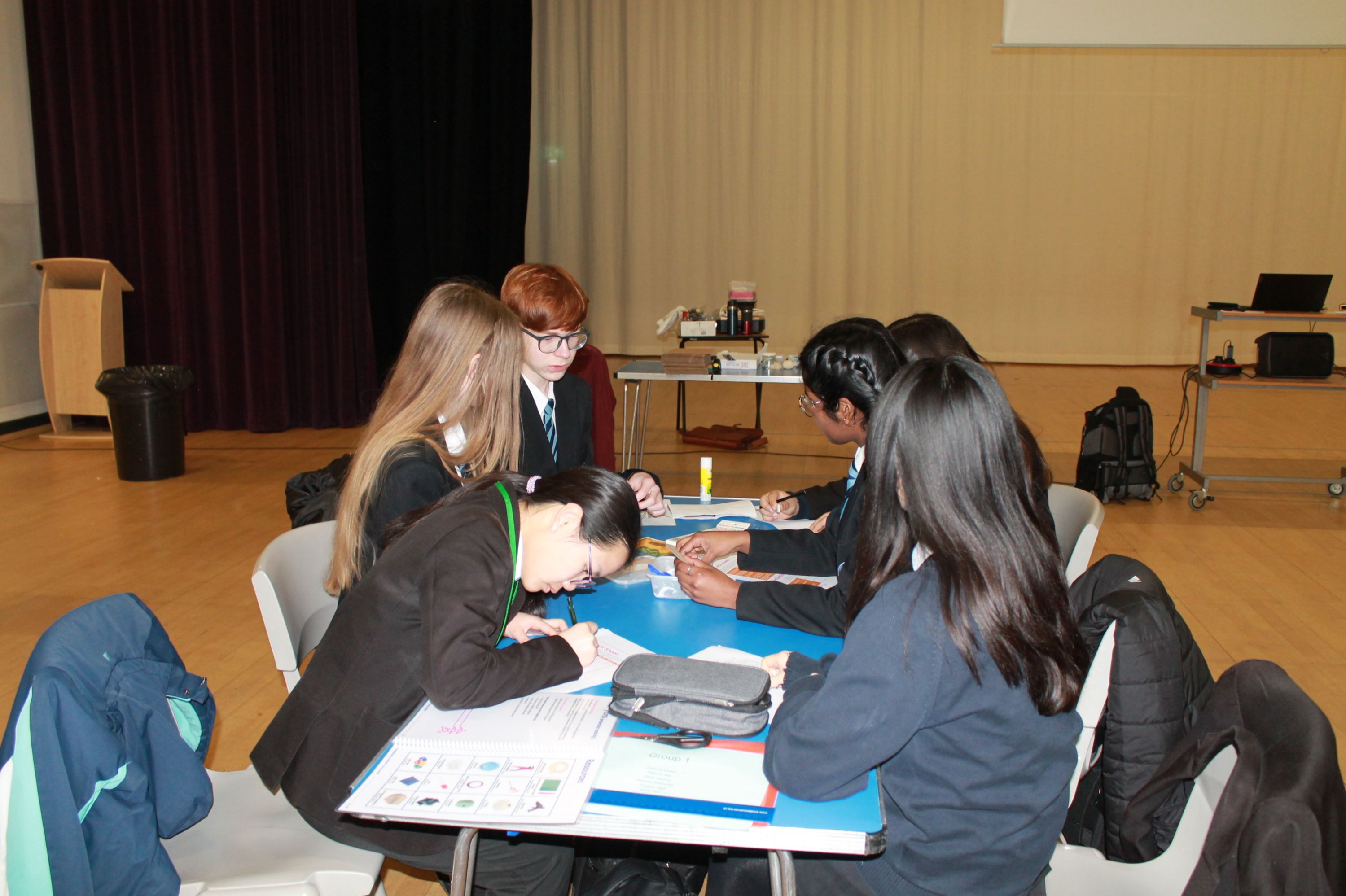 Photograph showing a group of five students seated around a blue table in a large room, engaged in writing or drawing activities. The setting includes a wooden floor, a podium in the background, and various items on a table behind the students