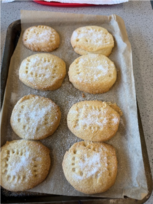 Photograph of eight freshly baked oval-shaped cookies arranged on a baking tray lined with parchment paper, lightly dusted with powdered sugar. Cookies vary slightly in size and have fork marks on top, indicating a homemade preparation.