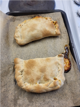 Photograph showing two baked pasties on a parchment-lined baking tray, placed on a stovetop. Pastries have golden-brown crusts with crimped edges, one slightly larger 