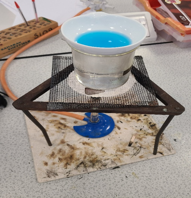 Photograph of a laboratory setup showing a blue liquid in a white ceramic crucible placed on a metal wire gauze supported by a tripod stand. The setup includes a Bunsen burner beneath the gauze, with various lab tools and equipment visible in the background, indicating a heating or chemical reaction experiment.