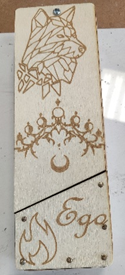 Photograph of a wooden rectangular object featuring laser-engraved designs including a geometric wolf head, a decorative chandelier, a crescent moon, and a flame symbol with the word "Ego