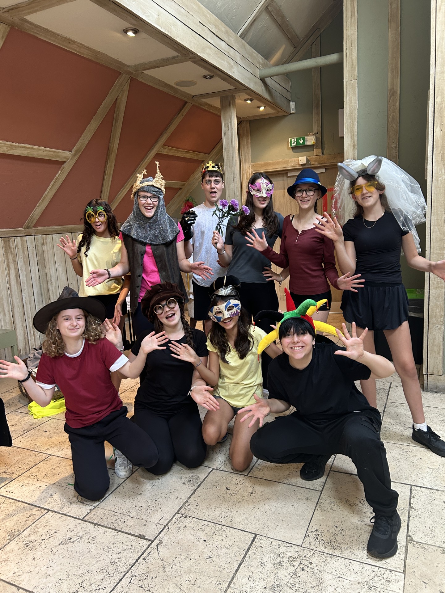 Photograph of a group of ten people dressed in various colourful costumes and accessories, posing indoors with wooden beams and a tiled floor. Costumes include a knight helmet, jester hat, masquerade masks, and a bridal veil, suggesting a playful or theatrical event.