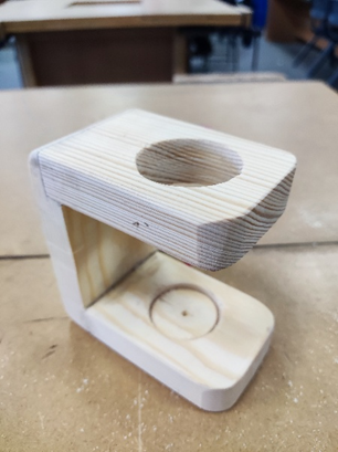 Photograph of a small wooden stand with a rectangular base and an elevated platform featuring a circular cutout. The stand appears handcrafted from light-coloured wood with visible grain patterns, designed to hold a round object securely in place.