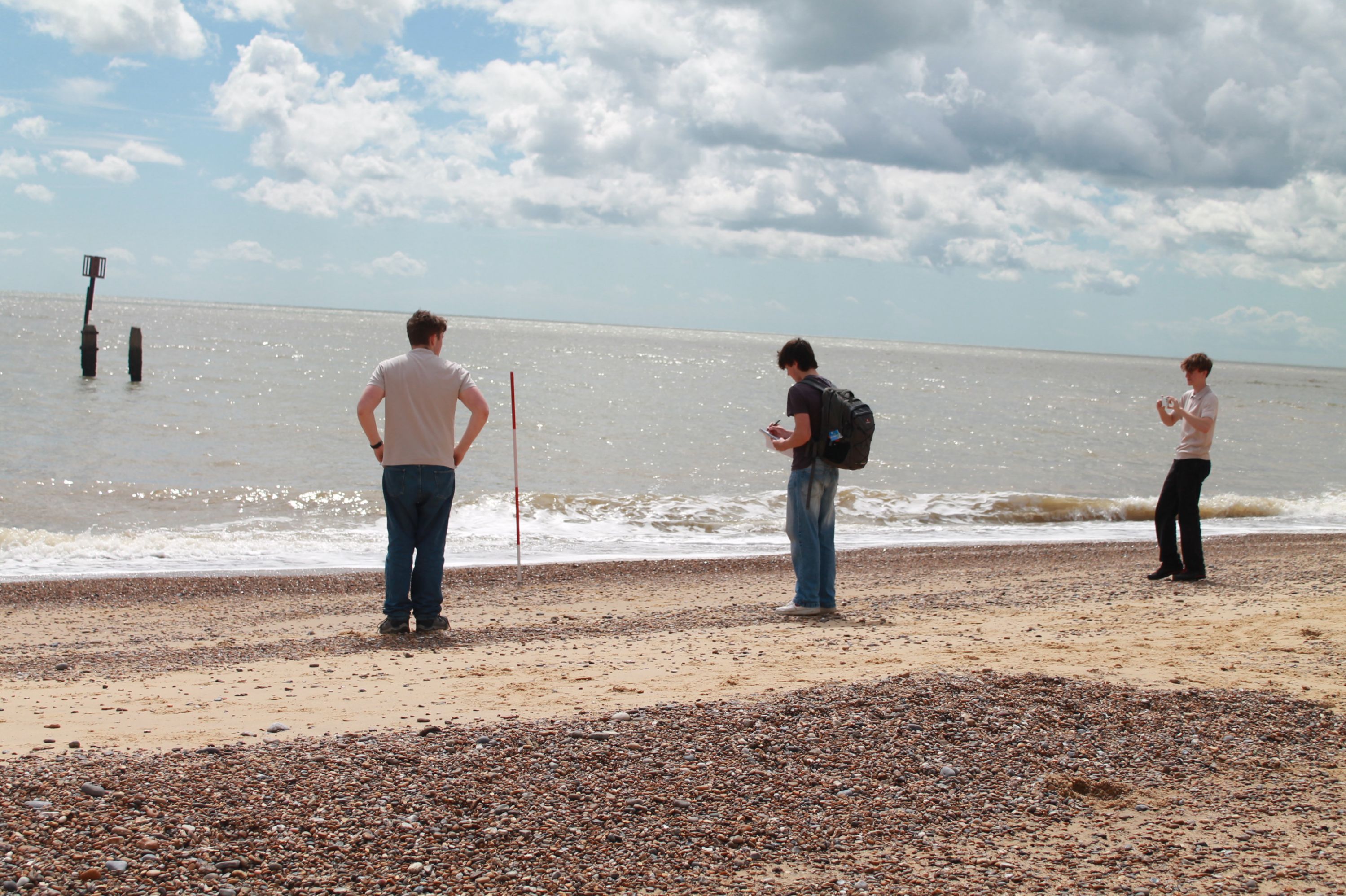 Photograph showing three people standing on a pebble and sand beach facing a calm sea under a partly cloudy sky. One person holds a red and white pole, another wears a backpack and looks at a device, and the third person appears to be taking a photo or video.