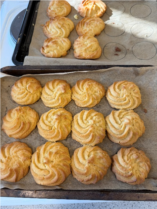 Photograph showing two baking trays with freshly baked, golden-brown swirl-shaped cookies on parchment paper. One tray contains a neat 3x4 grid of cookies, while the other has fewer cookies arranged irregularly, highlighting different stages of baking or preparation.