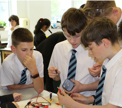 Photograph showing a group of schoolboys in white shirts and striped ties gathered around a table conducting a science experiment involving wires and a small device. The setting is a  laboratory, with students focused on hands-on learning and collaboration.