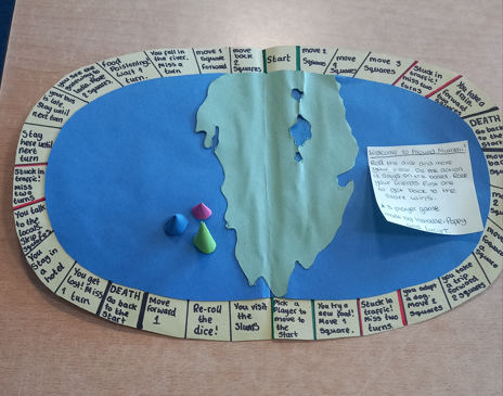 Photograph of a handmade board game featuring an oval track of beige spaces with handwritten instructions surrounding a blue and green central map. The game includes three coloured playing pieces, a dice, and a note explaining rules, with spaces indicating actions like moving squares, missing turns, and returning to start, highlighting a playful exploration theme.
