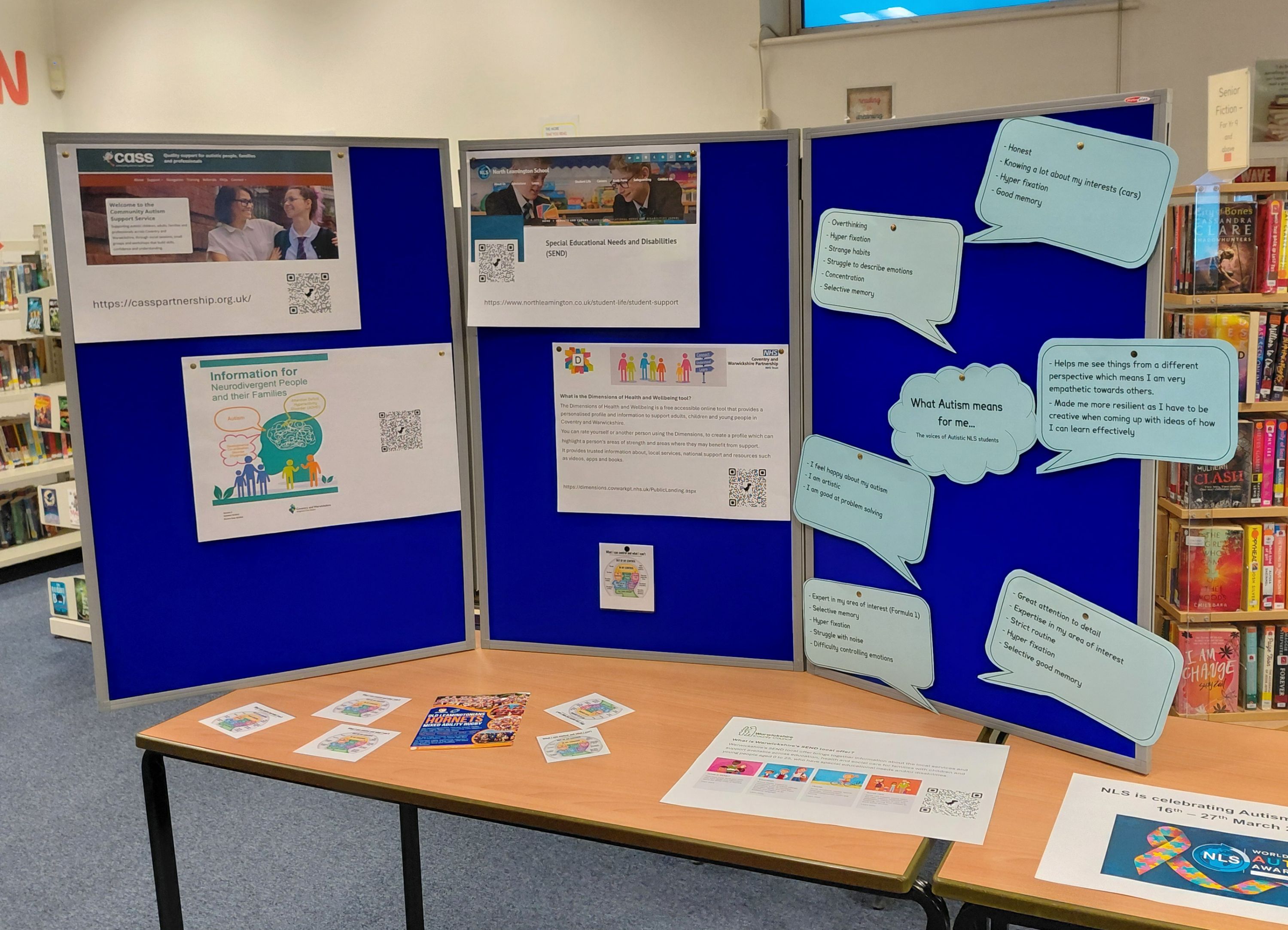 Photograph of an information display board set up in a library, featuring printed sheets and speech bubble cutouts on a blue background. Content focuses on autism awareness, including facts, support resources, and personal perspectives, with QR codes and colourful graphics