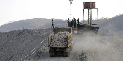hotograph showing a mining site with a large dump truck loaded with rocks driving up a dusty slope. Several workers wearing helmets stand near a tall metal structure, with hills visible in the background under a clear sk