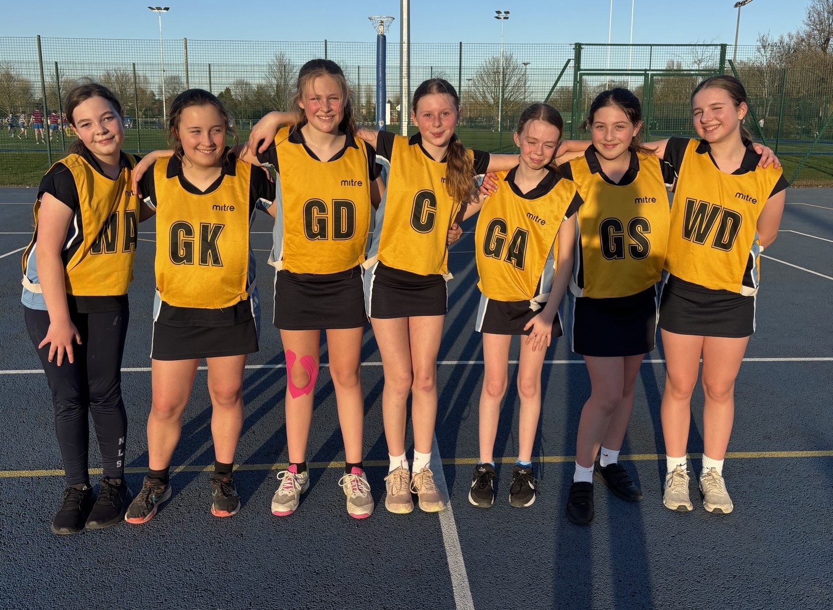 Photograph of a netball team standing side by side on an outdoor court, wearing yellow bibs with black position letters (GK, GD, C, GA, GS, WD) over black sportswear. The setting is during daylight with a clear sky, 