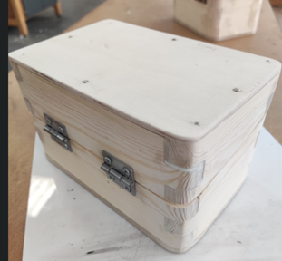 Photograph of a small rectangular wooden box with a hinged lid and metal clasps on the front side. Box features smooth, light-coloured wood with visible grain patterns and rounded edges, placed on a white surface