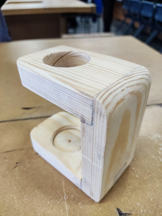 Photograph of a small wooden stand with two circular recesses, The stand features smooth, rounded edges and visible wood grain, placed on a workshop table with blurred background elemen