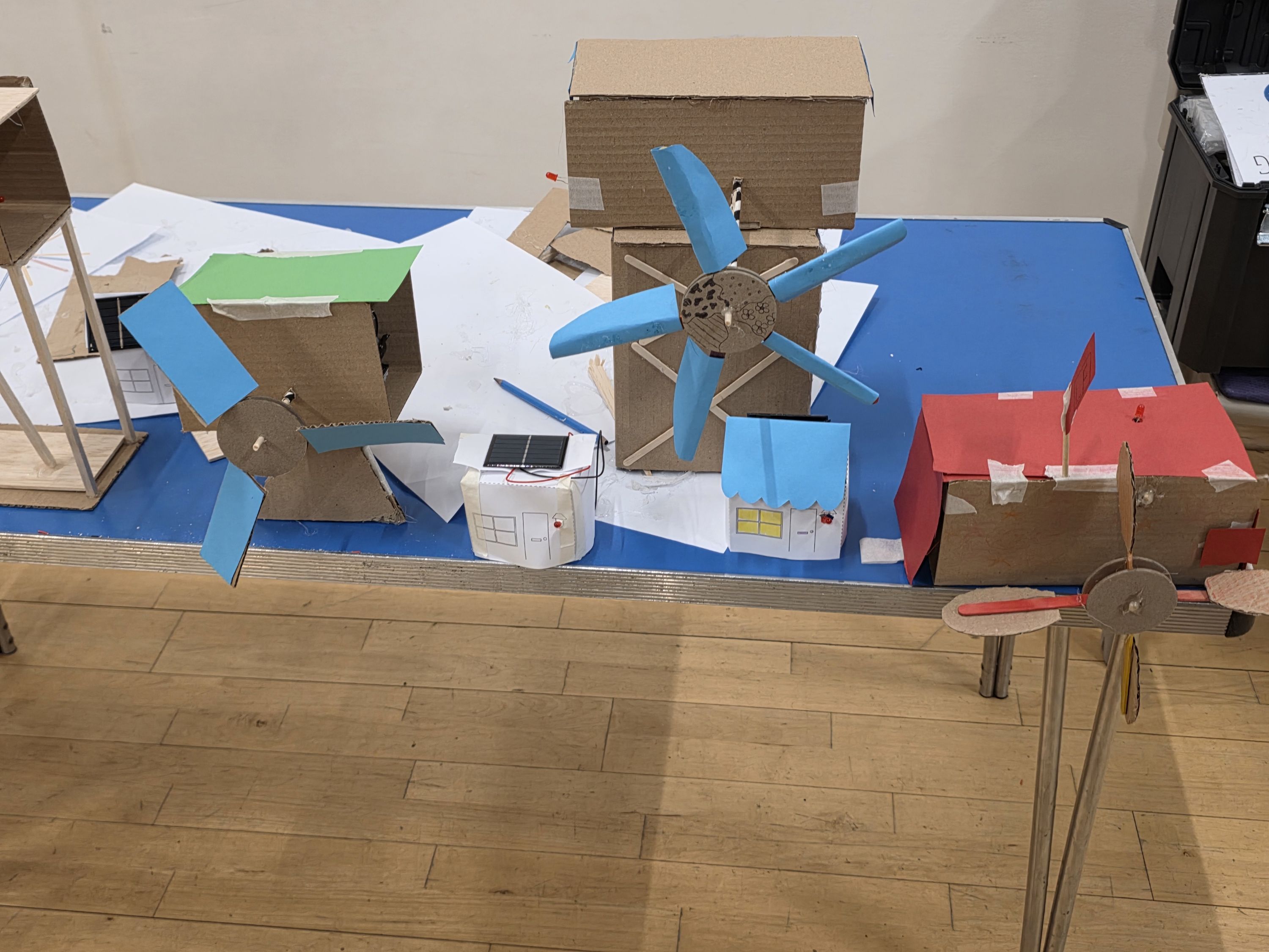 Photograph of a tabletop display featuring handmade cardboard models of wind turbines and small buildings, showcasing a renewable energy concept. Models include two wind turbines with blue blades, a red building with a propeller, and small houses with solar panels, arranged on a blue and white surface.