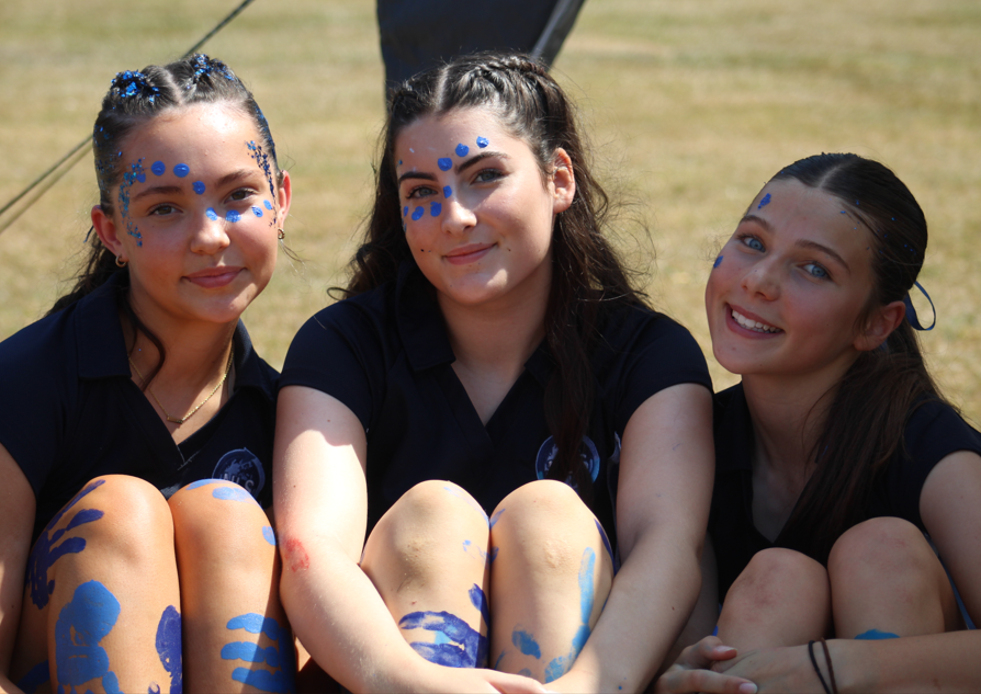 hotograph of three girls sitting outdoors on grass, each with blue face and body paint in patterns including dots and handprints. They wear dark shirts and smile at the camera, suggesting participation in a group or team event with a playful and creative atmosphere