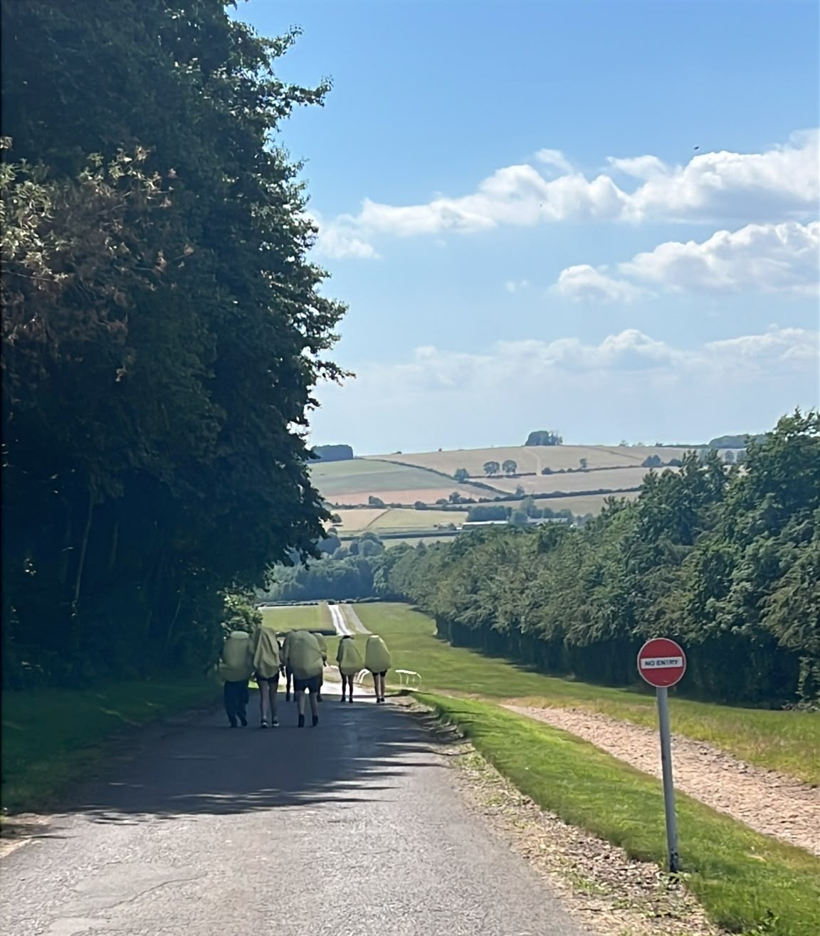 Photograph showing a group of people walking along a rural road bordered by trees and fields under a partly cloudy sky. A red and white "No Entry" sign is visible on the right side, indicating restricted access to the adjacent path.