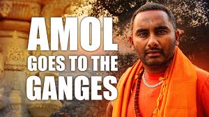 A photograph showing a person dressed in orange traditional attire with a blurred face, standing outdoors near an ornate structure. Bold white text overlay reads "AMOL GOES TO THE GANGES," indicating a theme related to a journey or visit to the Ganges River.