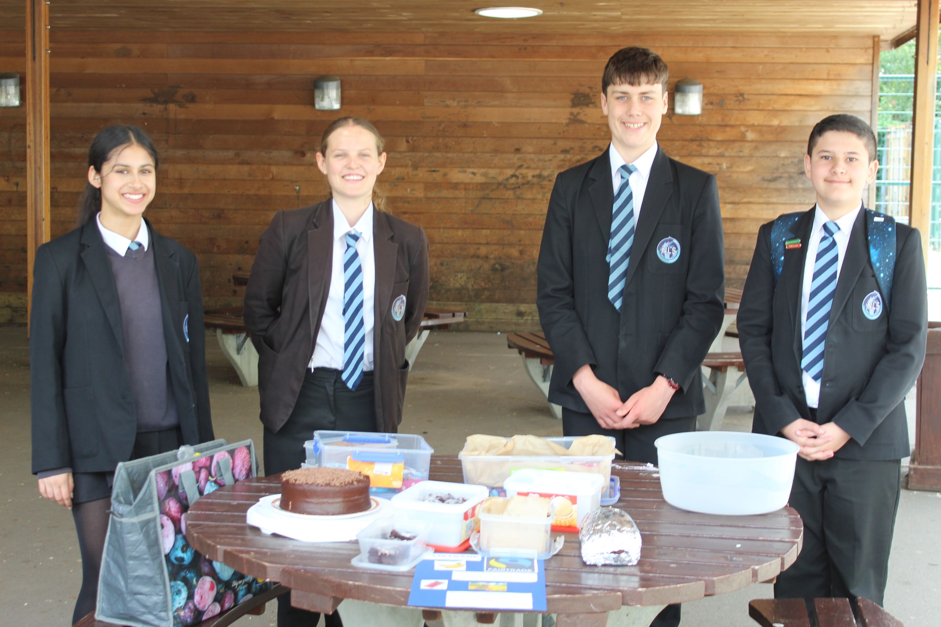 Photograph of four students in school uniforms standing behind a round table displaying various baked goods and containers, likely at a bake sale or fundraising event. Background features wooden panel walls and picnic benches, with students smiling and wearing blazers and striped ties.