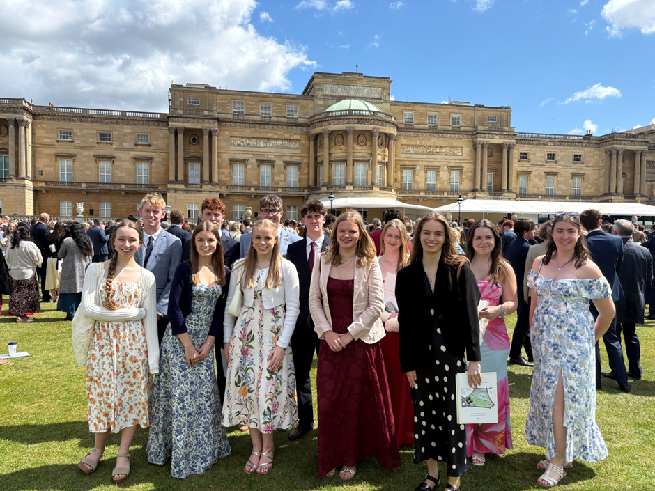 Photograph showing a group of young people dressed formally standing on grass in front of a large historic building under a partly cloudy sky at Buckingham Palace in a formal event or gathering, with individuals wearing suits and dresses in various colours and patterns, and a crowd visible in the background.