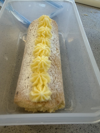 Swiss roll cake placed inside a transparent plastic container on a kitchen countertop. The cake is dusted with powdered sugar and decorated with a line of piped yellow cream along its top.