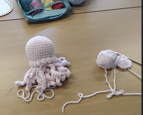 Photograph showing a handmade crocheted pink and white octopus with curly tentacles on a wooden table next to a partially crocheted piece and a ball of yarn with a crochet hook. A blue pouch with various yarns is visible in the background,