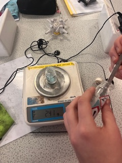 Photograph showing a digital weighing scale displaying 24.1 grams with a small glass container placed on it. Hands are holding a syringe near the scale, 