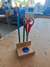 Photograph of a wooden desk organiser holding a blue pencil, a green pencil, and a pair of red scissors, placed on a workbench in a workshop setting. Background includes various tools, equipment, and a window, indicating a workspace environment.​​