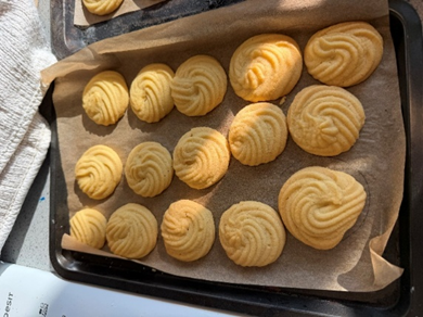 Photograph of a baking tray lined with parchment paper holding 15 round, piped cookies with a golden-brown colour. Cookies are arranged in four rows, showing slight variations in size and colour, indicating they have been freshly baked.