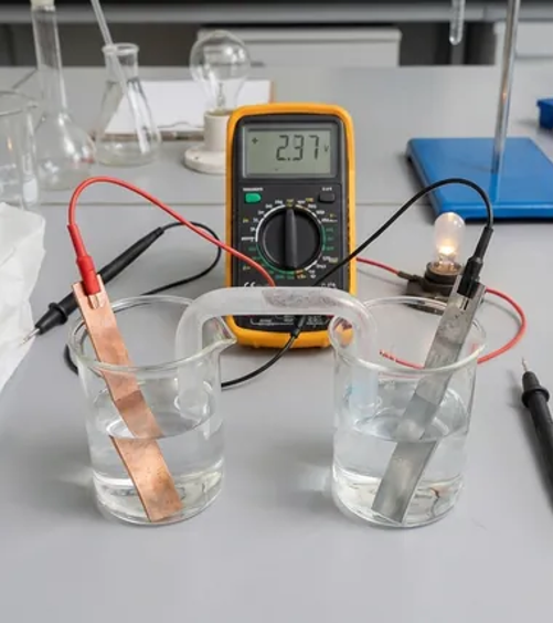 Photograph showing two experimental setups for measuring electrical properties in liquids, featuring multimeter and power supply connected to electrodes submerged in clear and blue solutions. Key elements include copper and steel electrodes, digital displays showing voltage and current, and visible light bulb indicating circuit activity.