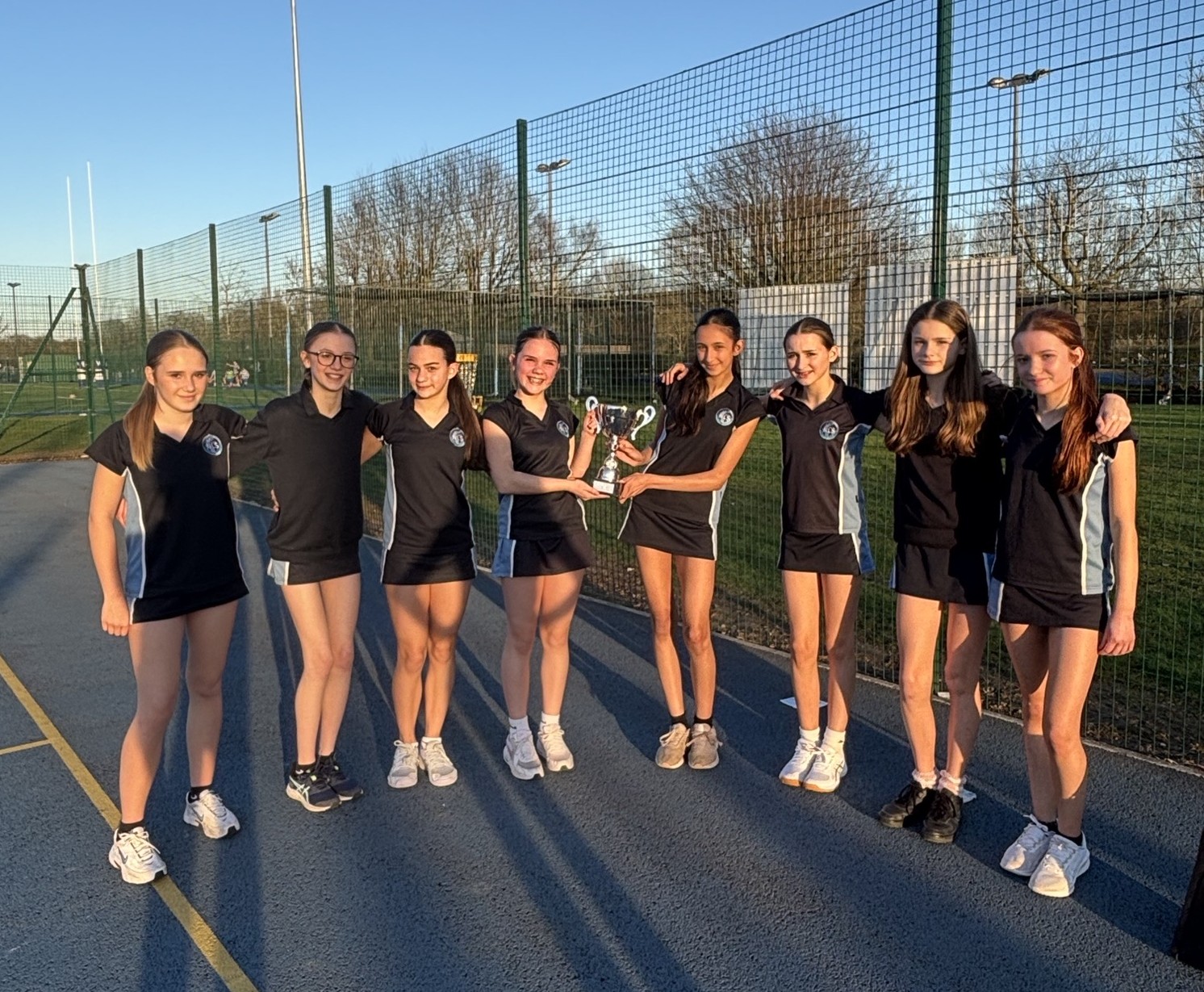 Photograph of a girls' netball team standing on an outdoor court with a netted fence and rugby posts in background, holding a trophy. Team members wear matching black sports uniforms with white accents, posing in sunlight with long shadows cast on the court.