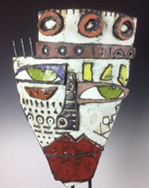    Title “another parade” by Kimmy Cantrell. Ceramic with glaze. A photograph of a colourful abstract mask featuring geometric shapes and bold patterns. The mask displays large green eyes, red lips, circular designs on the forehead, and a mix of white, black, red, and yellow colours creating a striking visual contrast.