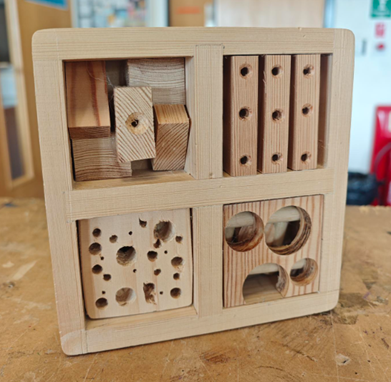 Photograph of a wooden insect hotel designed with multiple compartments featuring various hole sizes and shapes to provide shelter for different insect species. Sections include blocks with drilled holes, circular cutouts, and stacked wooden pieces, showcasing a natural, untreated wood finish on a workshop table.