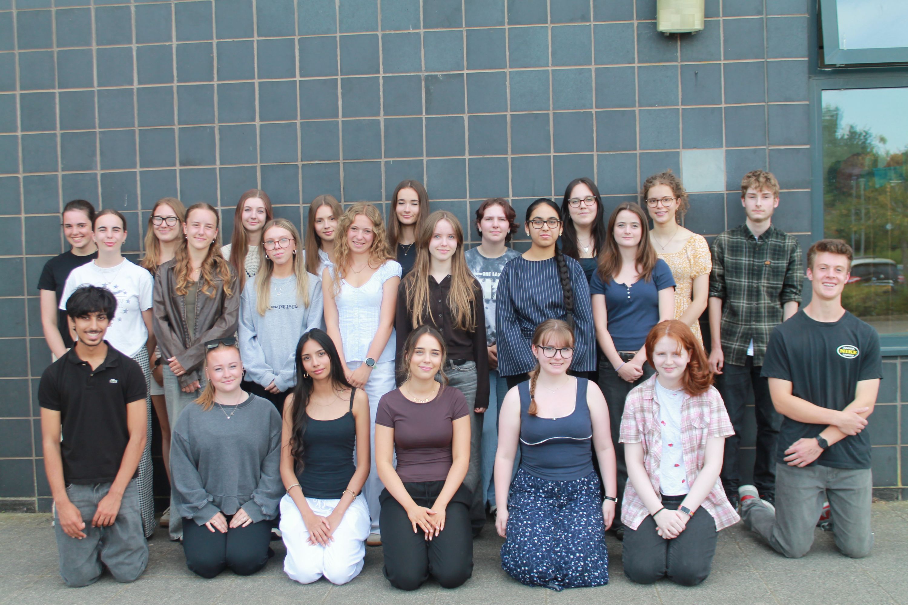 Photograph of a group of 23 young people posing outdoors against a tiled wall, arranged in two rows with some standing and others kneeling. The group displays diverse clothing styles and expressions, suggesting a casual or school-related gathering.