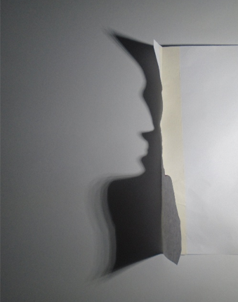Photograph showing a torn piece of white paper taped to a grey wall, casting a shadow resembling a human profile. The contrast between light and shadow creates an optical illusion highlighting the torn edge's shape.