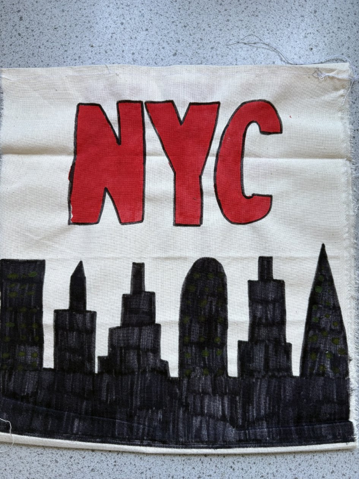 Photograph of a fabric square featuring a black silhouette of New York City skyline on the left and bold red letters "NYC" on the right. Design contrasts dark buildings against a light background, highlighting iconic skyscraper shapes and city abbreviation.