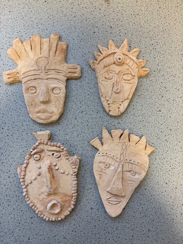 Photograph of four handcrafted clay masks arranged in a square on a speckled surface. Each mask features distinct facial expressions and decorative elements, including spikes, beads, and carved patterns, showcasing diverse artistic styles.