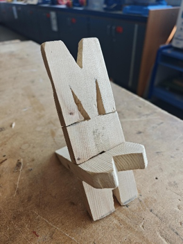 Photograph of a small wooden chair model shaped like the letter "M" with a backrest, seat, and legs, placed on a workbench. Background shows a workshop setting with cabinets and a blue chair, highlighting craftsmanship and design creativity.