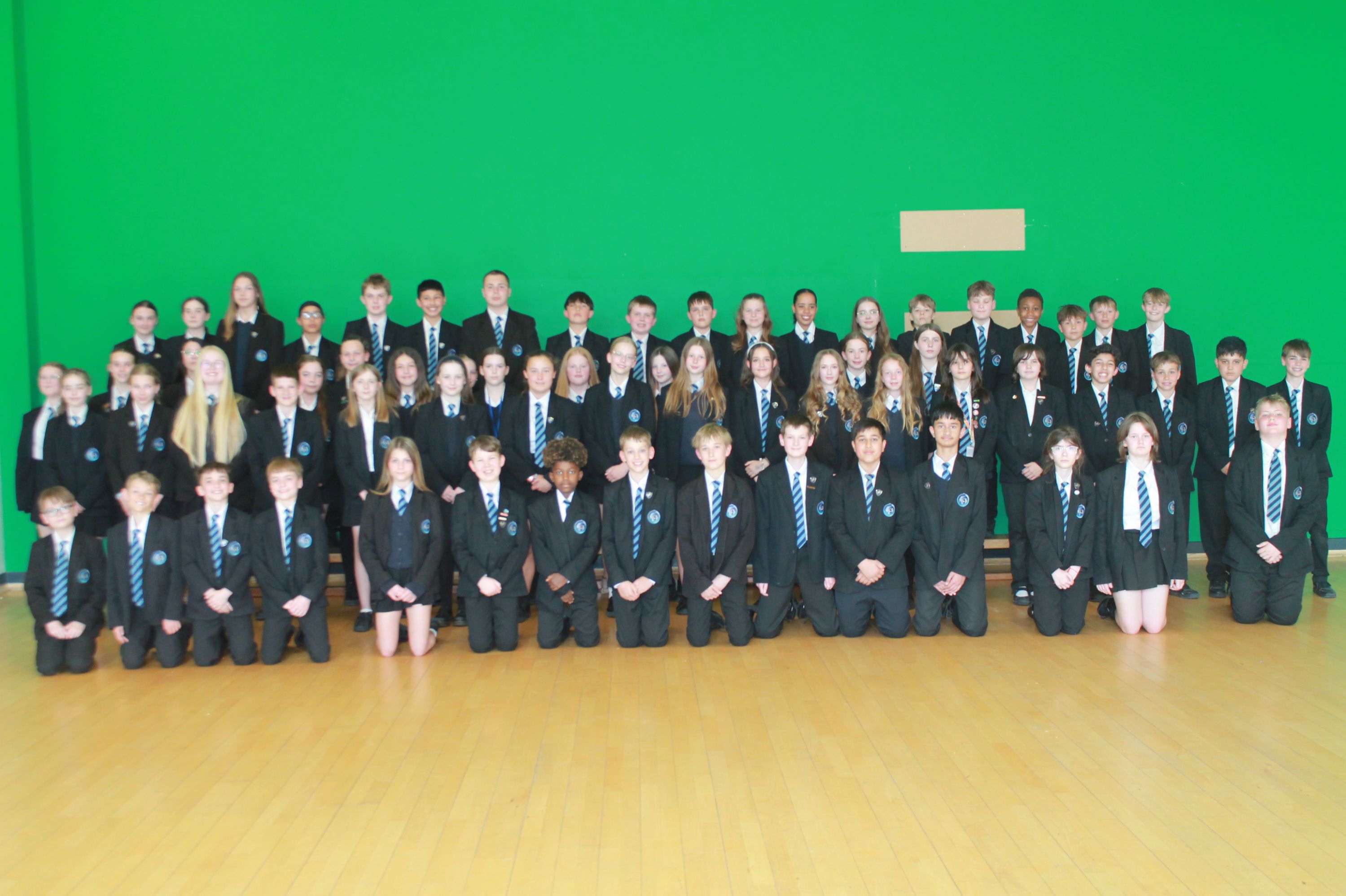 Photograph of a large group of school students in uniform posing indoors against a green wall. Students are arranged in three rows, with some standing and others kneeling, wearing black blazers, white shirts, and blue striped ties.