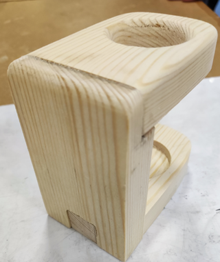 Photograph of a wooden object with a rectangular base and a vertical section featuring a rounded cutout at the top. The object displays visible wood grain and smooth edges, suggesting it may be a handcrafted item or a component for holding or supporting something.