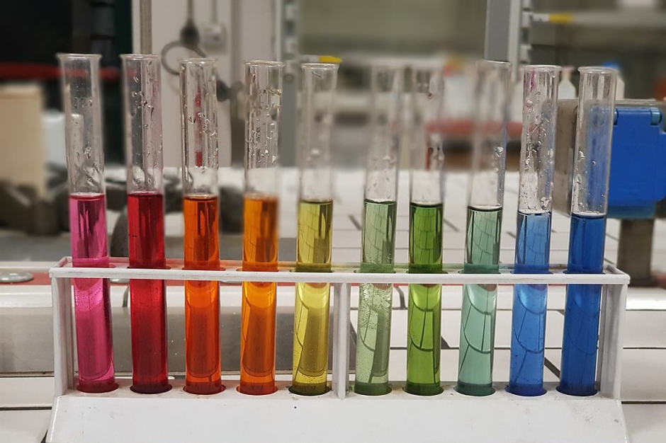 Photograph of ten test tubes arranged in a white rack, each containing liquid in a gradient of colours from pink, red, orange, yellow, green to blue. The setup appears to demonstrate a colour spectrum or pH indicator range, with clear glass tubes and a laboratory background.