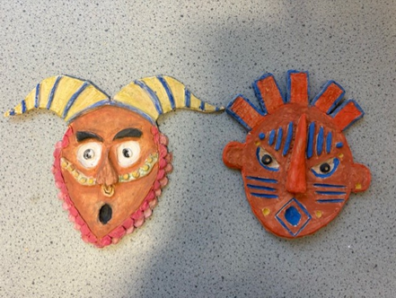 Photograph of two colorful handcrafted masks placed on a speckled surface, showcasing artistic facial designs and vibrant paint. One mask features a horned design with yellow and blue stripes, while the other has a red base with blue geometric patterns and protruding elements.